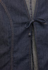 Vêtement en denim foncé avec une fermeture à nouer à l'avant. Le design comprend des coutures visibles dans une couleur contrastante et une surface légèrement texturée.