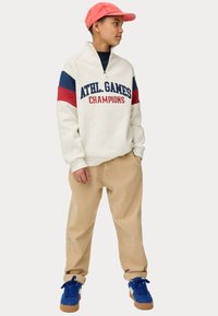 Sudadera blanca con rayas en azul marino y rojo, texto "CAMPEONES DE JUEGOS ATLÉTICOS", combinada con pantalones beiges y zapatillas azules con suelas marrones. Gorra rosa.