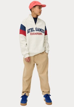 Witte sweatshirt met marineblauwe en rode strepen, tekst "ATHL. GAMES CHAMPIONS", gecombineerd met beige broek en blauwe sneakers met bruine zolen. Roze pet.