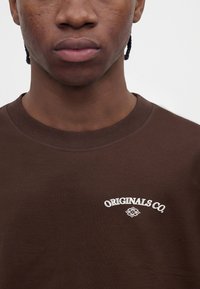 Gros plan d'une personne portant une chemise marron avec le texte blanc « ORIGINALS CO. » et un petit logo décoratif en dessous sur la poitrine.