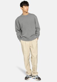 Grå crewneck sweatshirt med ribbede manchetter, parret med lysebeige løstsiddende bukser. Modellen står med hænderne i lommerne, iført atletiske sko.