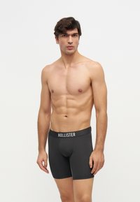 Jeune homme à la peau claire et aux cheveux foncés, portant un boxer noir Hollister, debout devant un fond blanc uni.