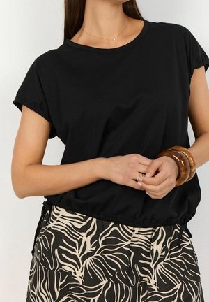 Kvinde iført en sort top med korte ærmer og et sort-beige skørt med abstrakt mønster, accessoriseret med træarmbånd og en delikat halskæde.