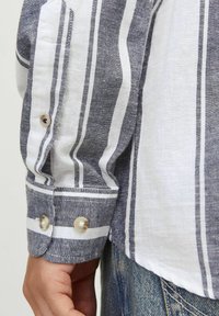Tessuto a righe blu e bianche con una texture di lino. La manica presenta due bottoni beige con una leggera lucentezza al polsino.