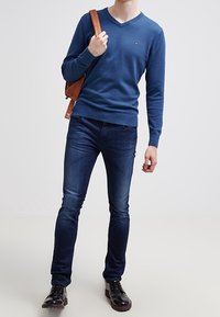 Niebieski sweter z dekoltem w serek, lekki dzianinowy; ciemne dżinsy o dopasowanym kroju; czarne buty; nosi brązową skórzaną torbę na ramię.