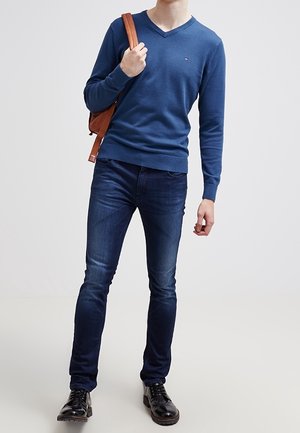 Modrý svetr s výstřihem do V, lehký pletený materiál; tmavé džíny s úzkým střihem; černé boty; nese hnědou koženou tašku přes rameno.