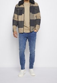 Chaqueta a cuadros en beige y azul marino con un cuello de piel sintética, combinada con una camiseta beige y jeans azul claro. Zapatillas blancas completan el conjunto.