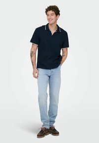 Jonge man draagt een donkerblauw poloshirt, lichtblauwe spijkerbroek en bruine leren schoenen, staat met één hand in de zak en glimlacht.