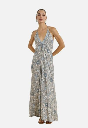 Mujer con un vestido sin mangas, de escote en V profundo y largo hasta el suelo, con un patrón floral en azul y beige, de pie con las manos en las caderas.