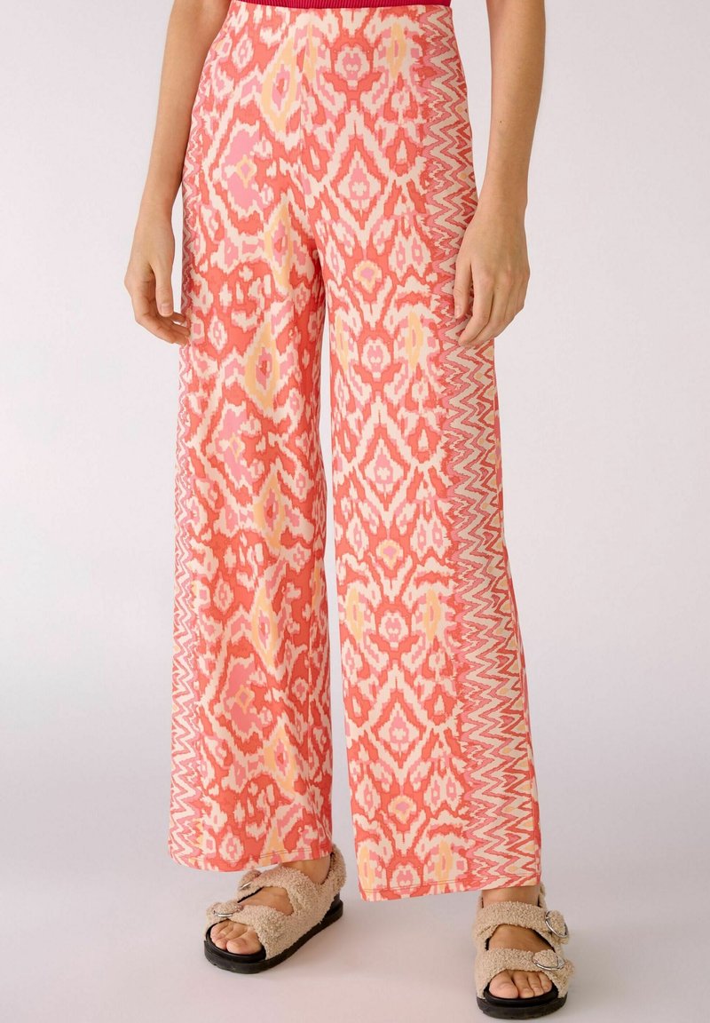 Pantalon à jambes larges avec un motif géométrique corail, rose et jaune, porté avec des sandales beiges duveteuses à boucles sur fond clair.