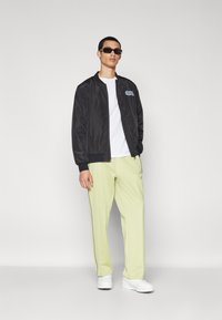 Black bomber jacket with ribbed cuffs, white t-shirt, and light green trousers. Accessories include dark sunglasses and white sneakers.
