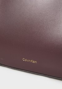Sac en cuir bordeaux avec une texture lisse, arborant le logo doré « Calvin Klein » en bas. Forme rectangulaire avec un design minimaliste.