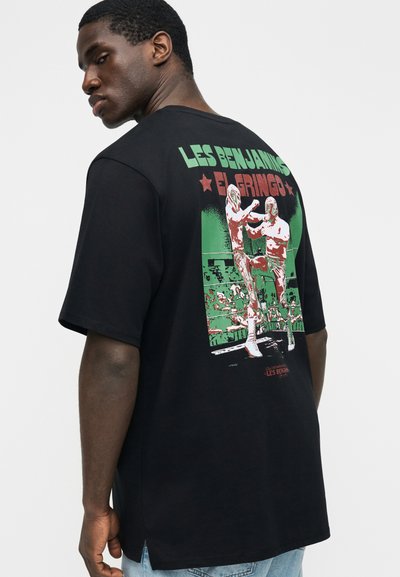 Άνδρας που φοράει μαύρο μπλουζάκι με σχέδιο δύο παλαιστών με μάσκες να παλεύουν σε ρινγκ και το κείμενο «Les Benjamins El Gringo» στην πλάτη.