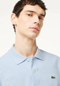 Ljusblå polo t-shirt med textilstruktur, två knappar på knappslån och en liten grön broderad krokodil-logga på bröstet.