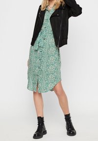 Abito verde a fiori con bottoni, maniche corte, abbinato a una giacca in suede nera e stivaletti neri con lacci. I dettagli notevoli includono un colletto e spacchi laterali.