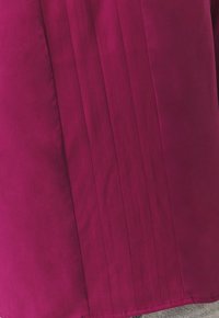Blusa magenta con una consistenza liscia, caratterizzata da pieghe verticali su un lato e un orlo curvo, realizzata in tessuto leggero.
