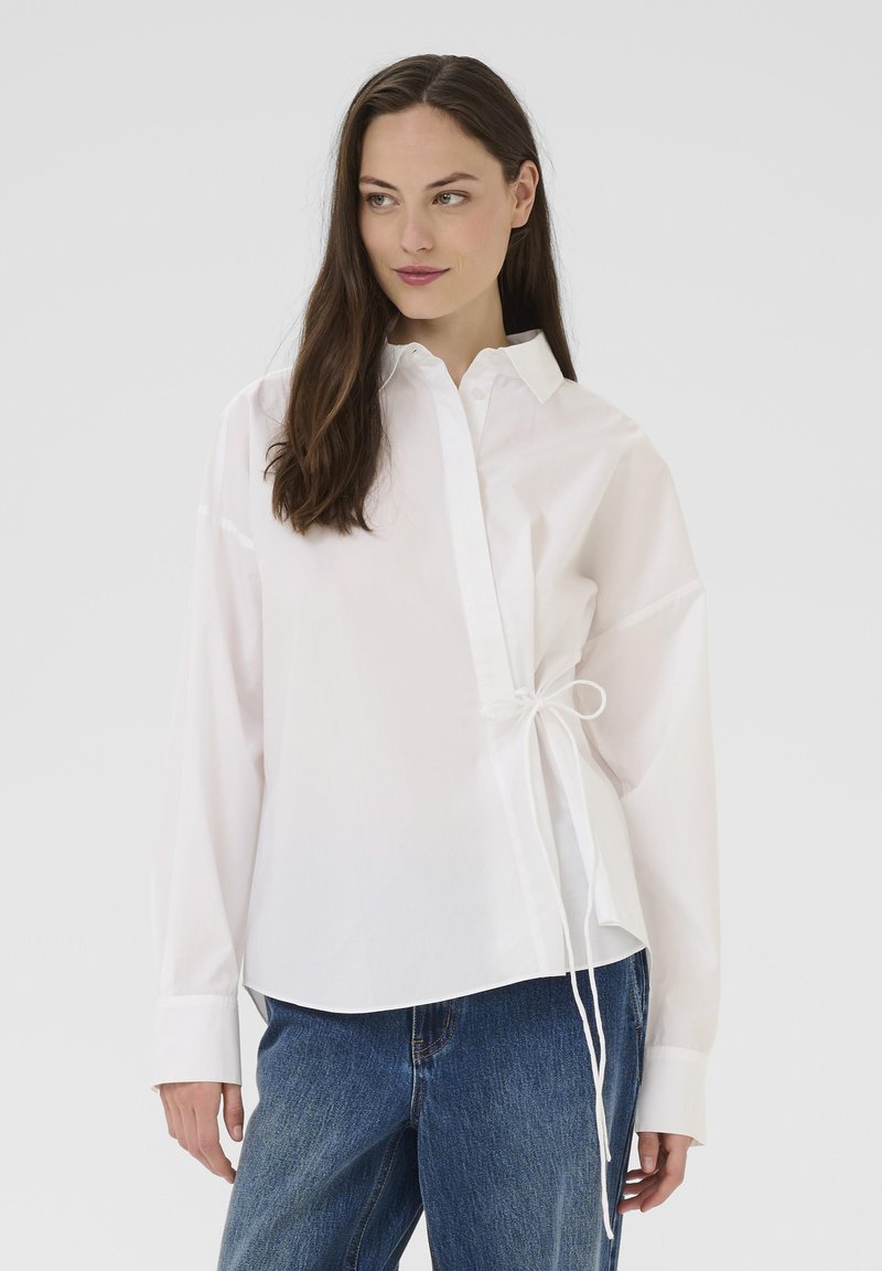 White long-sleeve shirt made of smooth fabric, featuring a button-up front, collar, and a tied accent detail on the side.