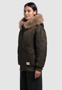 Geacă puffer verde oliv cu glugă căptușită cu blană, având buzunare cu fermoar și capse, închidere cu fermoar și manșete cu nervuri. Textură netedă, căptușită.