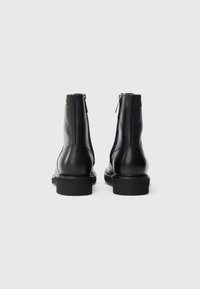 Une paire de bottines noires en cuir avec des talons carrés et des fermetures éclair à l'arrière, présentée de l'arrière sur un fond clair.