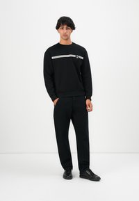 Young man standing wearing a black sweatshirt with white stripes, black pants, and black sneakers against a plain white background.