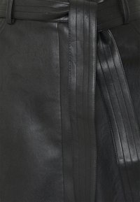 Svart läderplagg med bredt bälte som har stickade vertikala linjer och bältesöglor, vilket visar textur och sömdetaljer.