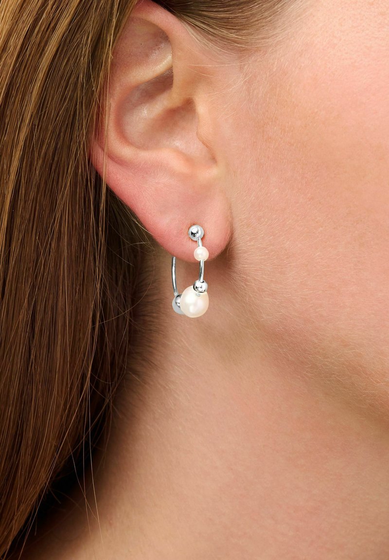 Orecchini a cerchio in argento con una finitura lucida, caratterizzati da due perle bianche e accenti sferici, indossati all'orecchio di un modello.