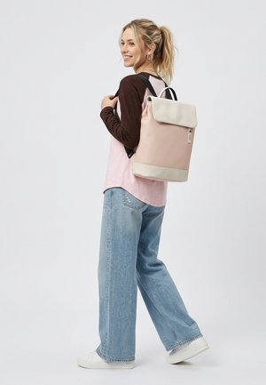 Jonge vrouw met blond paardenstaart, gekleed in een lichtroze en donkerbruine shirt, losse blauwe jeans, witte sneakers en een lichtroze rugzak.