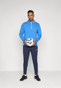 Haut de sport à manches longues bleu avec fermeture éclair et logo, jogging bleu marine, chaussures de foot noires, tenant un ballon de foot blanc à motifs.