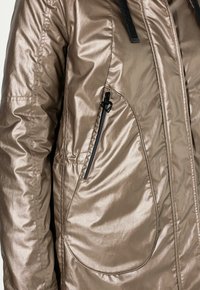 Chaqueta marrón metálica con una textura suave, que cuenta con un bolsillo con cremallera y detalles en negro. Incluye una capucha con cordón y detalles cosidos.
