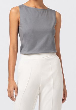 Haut gris clair sans manches associé à un pantalon blanc taille haute porté par une personne debout sur un fond uni.