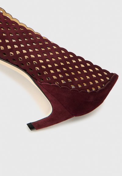 Scarpa alta in suede bordeaux con motivo traforato, bordi scallop e tacco lucido nero. Presenta una superficie testurizzata.