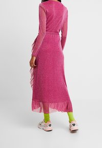 Largo vestido rosa con purpurina, mangas transparentes, detalle de volantes en el costado y un dobladillo fluido. Lleva calcetas verdes y zapatillas multicolor.