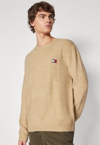 Pull en tricot beige avec des poignets et un ourlet côtelés. Présente un motif en torsades sur le devant et un petit patch logo sur la poitrine gauche.