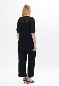 Maglietta nera a maglia con texture semi-trasparente e maniche a sbuffo; abbinata a pantaloni neri a gamba larga e sandali con tacco neri. Vista posteriore.