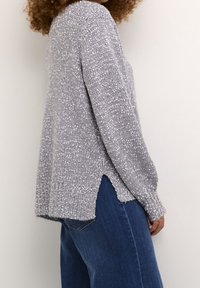 Pull tricoté gris clair à coupe ample, avec une fente latérale, des poignets côtelés et une finition texturée. Associé à un jean en denim bleu.