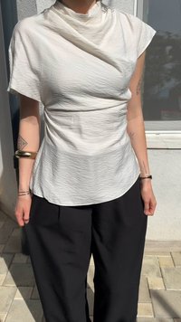 White, crinkled fabric top with a draped neckline and short sleeves, paired with loose black trousers. Visible tattoos on arms.