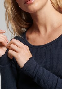 Dunkelblaues Strickoberteil mit gerippter Textur, rundem Halsausschnitt und ausgefransten Bündchen. Hände berühren den Stoff in der Nähe des Halsausschnitts.