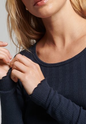 Granatowy sweter z dzianiny o prążkowanej fakturze, okrągłym dekolcie i postrzępionych mankietach. Ręce dotykają materiału w okolicy dekoltu.