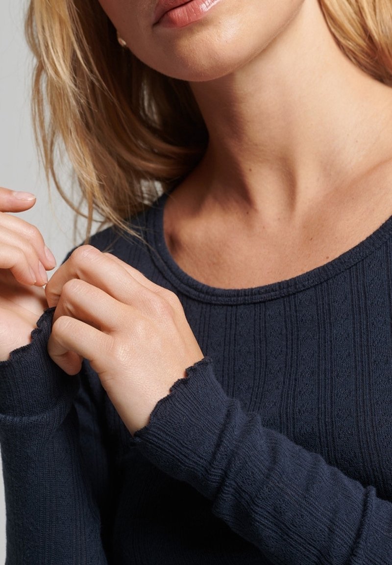 Dunkelblaues Strickoberteil mit gerippter Textur, rundem Halsausschnitt und ausgefransten Bündchen. Hände berühren den Stoff in der Nähe des Halsausschnitts.