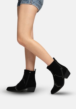 Benedenpartijen met spijkershorts en zwarte suède enkellaarzen met lage hakken en decoratieve zilveren studs, tegen een effen lichte achtergrond.