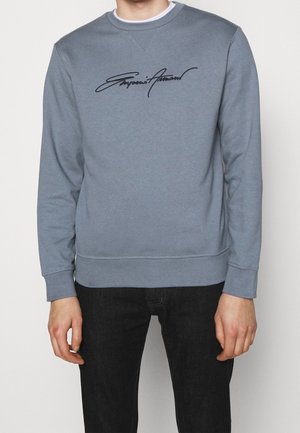 Personne portant un sweat-shirt bleu clair avec le logo script "Emporio Armani" sur la poitrine et un pantalon noir, debout devant un fond uni.