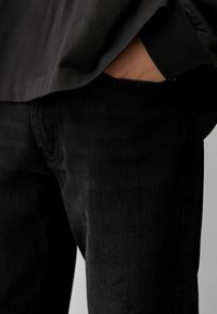 Osoba koja nosi crne traperice i crnu majicu dugih rukava, s desnom rukom u prednjem džepu.