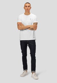T-shirt en coton blanc avec un petit imprimé de visage souriant noir, associé à un jean en denim foncé et des baskets blanches. Design simple et coupe décontractée.