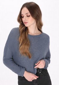 Pull en tricot gris avec une finition texturée, encolure ronde et manches longues, associé à un jean noir taille haute.