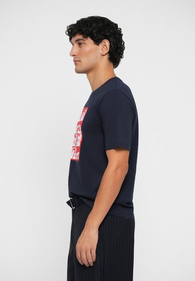 Camiseta de algodón azul marino con cuello redondo, con un estampado gráfico rojo. Manga corta, ajuste slim, combinada con pantalones de rayas.