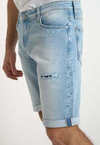 Pantalones cortos de mezclilla azul claro con detalles desgastados, dobladillos enrollados y un acento multicolor en el dobladillo. Diseño de cinco bolsillos con cierre de botón.