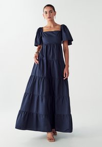 Navy maxi-mekko, jossa on neliönmuotoinen pääntie ja puffihihat, kerroksellisia röyhelöitä ja sileä kankaan pinta. Yksinkertainen, elegantti muotoilu.