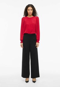 Blusa roja de manga larga con tejido texturizado, combinada con pantalones anchos de talle alto en negro. Tacones altos negros con acentos dorados.