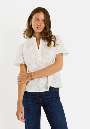 Mujer con cabello castaño ondulado que lleva una blusa blanca de manga corta con bordado floral y vaqueros azul oscuro, posando con los brazos cruzados.