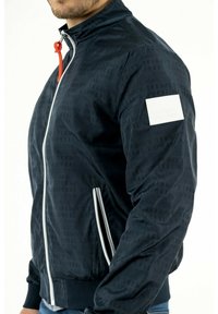 Giacca bomber navy con motivo logo sottile, dotata di una toppa bianca sul braccio, zip completa e zip bianche a contrasto. Texture morbida.